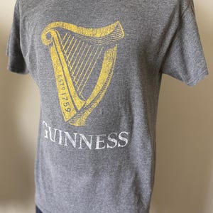 以下が含まれることがあります： 黄色いハープのデザイン、テキスト「ESTD 1759」、白い文字で「GUINNESS」がプリントされたグレーのTシャツ。半袖のTシャツは柔らかい素材で作られています。