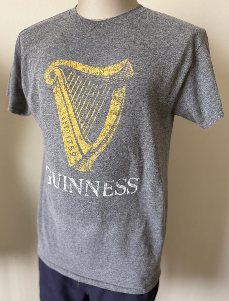 以下が含まれることがあります： 黄色いハープのデザイン、テキスト「ESTD 1759」、白い文字で「GUINNESS」がプリントされたグレーのTシャツ。半袖のTシャツは柔らかい素材で作られています。