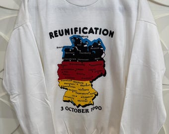 Puede incluir: Sudadera blanca con un gráfico del mapa alemán en los colores de la bandera alemana. La palabra "REUNIFICATION" está impresa encima del mapa, con "3 OCTOBER 1990" debajo. La sudadera tiene cuello redondo y mangas largas.