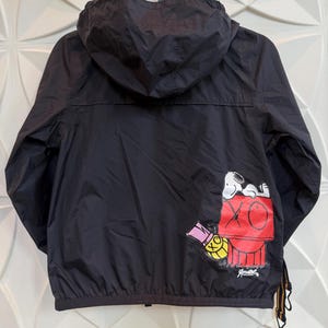 May include: Black hooded jacket featuring a Snoopy graphic on the back. The graphic includes Snoopy on top of a red doghouse with the text "XO" and "Andre". The jacket has a zipper closure and a drawstring at the bottom.
