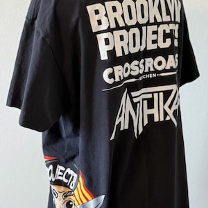 May include: Black t-shirt featuring the text "BROOKLYN PROJECTS CROSSROADS KITCHEN ANTHIK" in white. A cartoon graphic with a chef holding a knife is on the side. The shirt is displayed on a mannequin.