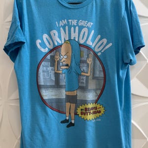 May include: Light blue t-shirt featuring a cartoon character with the text "I AM THE GREAT CORNHOLIO!". The character is inside a red and white circle. The shirt also has the text "BEAVIS AND BUTT-HEAD".