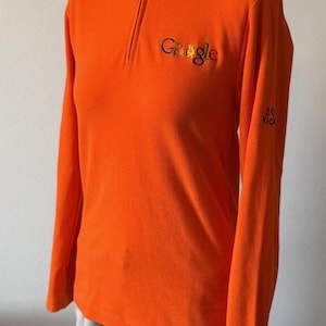 May include: Bright orange long-sleeve pullover with a mock neck and a quarter-zip closure. The Google logo is embroidered on the chest. The sleeve has the text "20 Kick". The shirt is displayed on a mannequin.