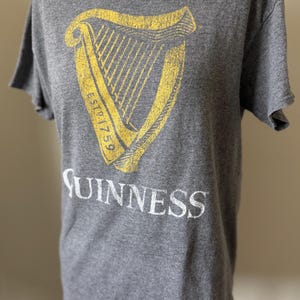 以下が含まれることがあります： グレーのTシャツ。黄色と白のギネスのロゴがプリントされています。ロゴにはハープと「ESTD 1759」の文字が含まれています。「Guinness」の文字がロゴの下に白で印刷されています。