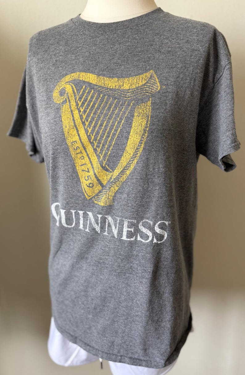 以下が含まれることがあります： グレーのTシャツ。黄色と白のギネスのロゴがプリントされています。ロゴにはハープと「ESTD 1759」の文字が含まれています。「Guinness」の文字がロゴの下に白で印刷されています。