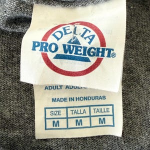 以下が含まれることがあります： グレーのTシャツのクローズアップ。白いタグには、赤と青の「DELTA PRO WEIGHT」のロゴが入っています。その下に「ADULT」と「MADE IN HONDURAS」とサイズ表があり、「M」が表示されています。