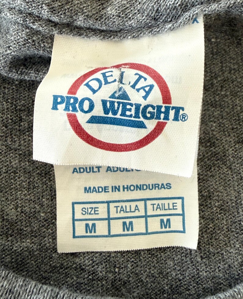 以下が含まれることがあります： グレーのTシャツのクローズアップ。白いタグには、赤と青の「DELTA PRO WEIGHT」のロゴが入っています。その下に「ADULT」と「MADE IN HONDURAS」とサイズ表があり、「M」が表示されています。