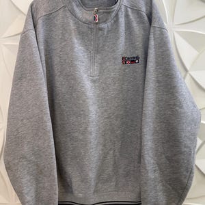 May include: A grey sweatshirt with a quarter-zip and the "Carnival" logo. The sweatshirt features a ribbed collar, cuffs, and hem with navy blue stripes. The long sleeves extend to the wrists.