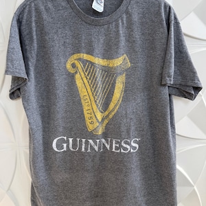 以下が含まれることがあります： ギネスのロゴが入ったグレーのTシャツ。ロゴは金色のハープで、下に白で「GUINNESS」と書かれています。Tシャツはクルーネックで半袖です。