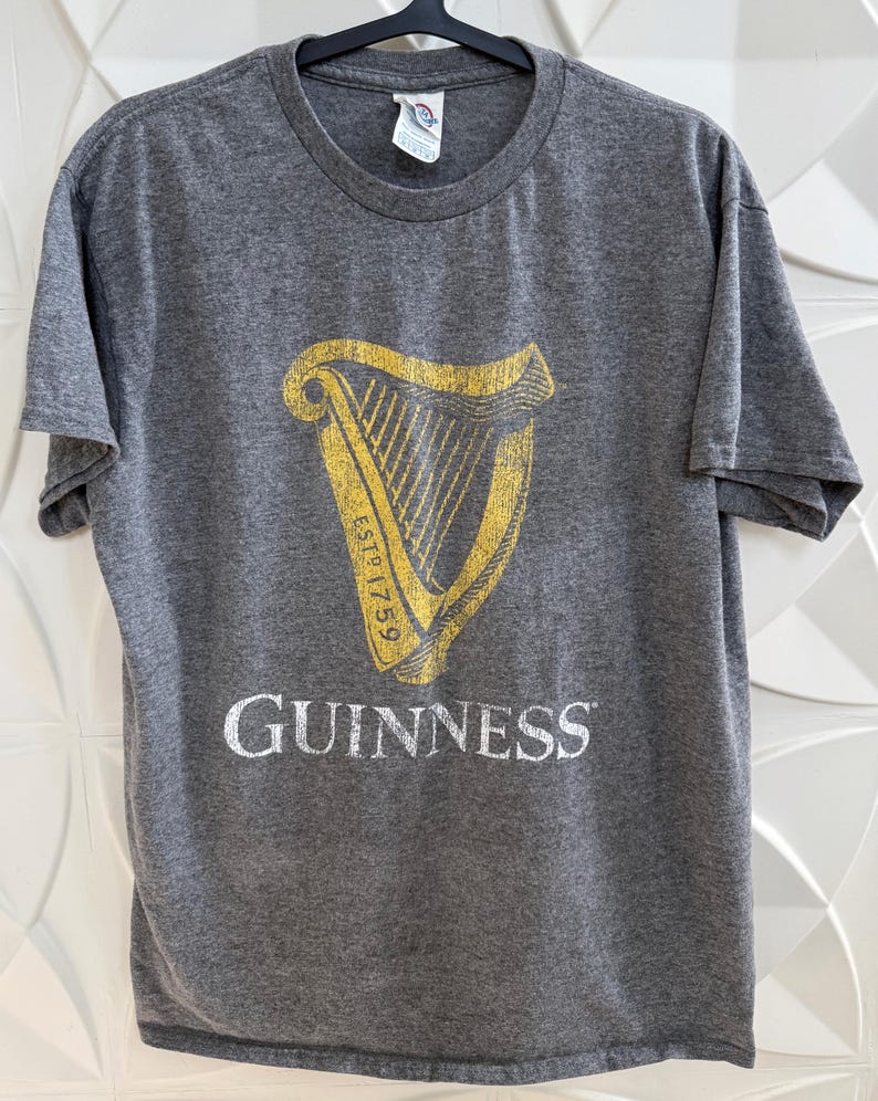 以下が含まれることがあります： ギネスのロゴが入ったグレーのTシャツ。ロゴは金色のハープで、下に白で「GUINNESS」と書かれています。Tシャツはクルーネックで半袖です。