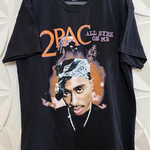 Könnte beinhalten: Schwarzes T-Shirt mit einem Porträt von 2Pac, dem Text "2PAC" in Orange und "ALL EYEZ ON ME" in Lila. Das Shirt hat einen Rundhalsausschnitt und kurze Ärmel. Das Bild zeigt ein Porträt von 2Pac mit einem Bandana.
