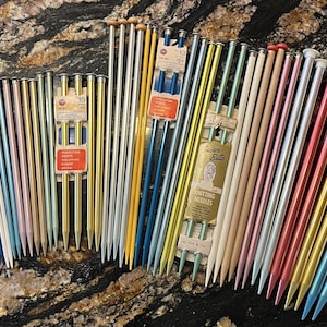 May include: A collection of vintage knitting needles in various colors, including silver, gold, blue, pink, and green. The needles are arranged in a fan shape, with the tips pointing towards the right. The needles are labeled with the size and brand "Susan Bates".