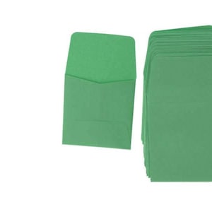 Puede incluir: Una pila de 25 sobres verdes, con un sobre colocado plano delante de la pila. Los sobres están hechos de papel texturizado.