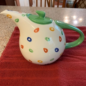 May include: White ceramic teapot with a green handle, lid, and spout. The teapot is decorated with colorful, ring-shaped designs in orange, blue, yellow, and green. The teapot is sitting on a red towel.