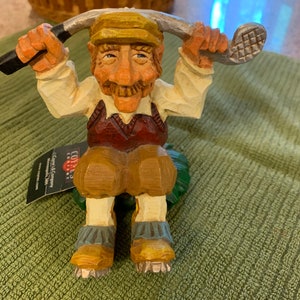 May include: A wooden figurine of a golfer wearing a brown vest, white shirt, and a yellow hat. The golfer is holding a golf club above his head. The figurine is sitting on a green base.
