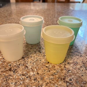 May include: Four small plastic containers with lids. The containers are white, light blue, yellow, and green.
