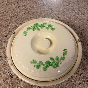 May include: Cream-colored ceramic casserole dish with a lid. The lid features green floral designs and a small knob. The dish has a round shape and a slightly distressed appearance, suggesting it is vintage.