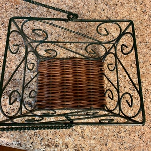 May include: A green metal hanging basket with a woven brown wicker insert. The basket has a decorative scrolled design.
