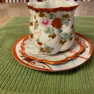 May include: A white porcelain teacup and saucer set with a floral design. The cup has a scalloped rim and the saucer has a scalloped edge. The floral design features pink, red, and green flowers and leaves.