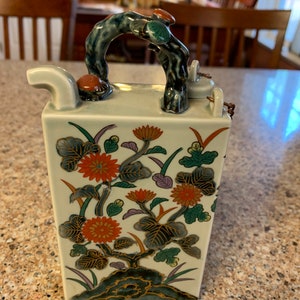May include: A ceramic teapot with a floral design. The teapot has a unique handle shaped like a tree branch. The teapot is decorated with red, orange, green, and gold flowers and leaves.