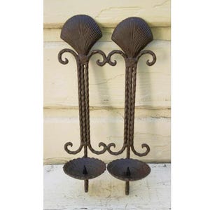 May include: A pair of wrought iron wall mounted candle holders with a decorative fan design at the top. The candle holders are dark brown and have a rustic look.