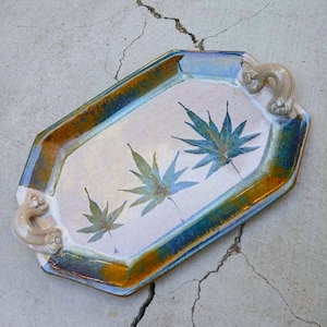 May include: Ceramic serving tray with a rectangular shape and decorative handles. The tray features a white interior with three blue-green leaf designs. The rim has a gradient of blue, brown, and gold. The handles are a light beige color.