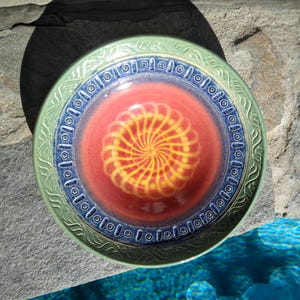 May include: A handmade ceramic bowl with a spiral design. The bowl features a red center with a yellow spiral, surrounded by a blue band and a green rim. The bowl is likely used for serving food or as a decorative piece.