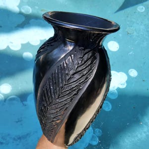 May include: A black ceramic vase with a glossy finish. The vase features a carved leaf design on one side and a flared rim. The vase is held against a blue background.