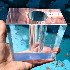 May include: A clear, rectangular crystal vase with a cylindrical opening. The vase is held in a hand, showcasing its geometric design and transparency. The vase is likely for floral arrangements or decorative purposes.