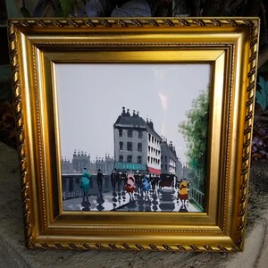 May include: A framed painting depicting a European street scene with figures and buildings. The artwork features a gold ornate frame. The painting shows a rainy day with reflections on the street. The color palette includes gray, black, and touches of color.