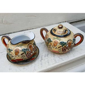May include: A vintage Japanese porcelain tea set featuring a creamer and a covered sugar bowl. The cream-colored pieces are adorned with detailed illustrations of figures, trees, and gold accents. The handles and rims are brown and red.