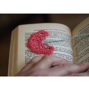 以下が含まれることがあります： 花の形をしたピンクのクロシェのブックマークのクローズアップ。ブックマークは開いた本の上に置かれ、手はページを持っています。