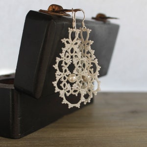 Peut inclure: Une paire de boucles d'oreilles en dentelle blanche avec des accents de perles, présentées dans une boîte en bois noir.
