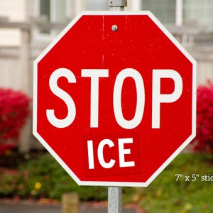 May include: A red octagonal stop sign with white text reading STOP and a rectangular sticker below it reading ICE. The sign is mounted on a metal pole. Text on the image indicates the sticker dimensions are 7 inches by 5 inches.