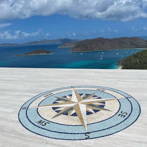 May include: A white tiled patio with a compass mosaic in the center. The compass is surrounded by a blue and white border. The compass points north, south, east, and west. The background shows a view of a blue ocean and green islands.