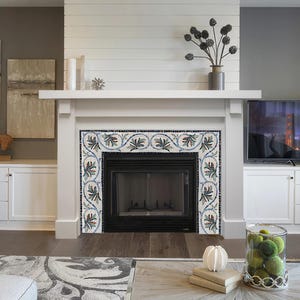 Op de afbeelding: Een witte schouw met een decoratieve tegelrand met een bloemmotief in tinten blauw, groen en wit. De open haard heeft een zwart metalen rooster en een witte omlijsting.