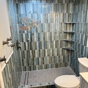 May include: A bathroom shower with blue and green tile, a shower head, and a sea turtle design. The shower has a built-in bench and a drain in the floor. There is a toilet in the background.
