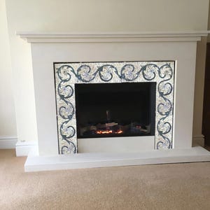 May include: White fireplace with a decorative mosaic tile border around the opening. The fireplace has a black metal firebox with a glowing fire inside.
