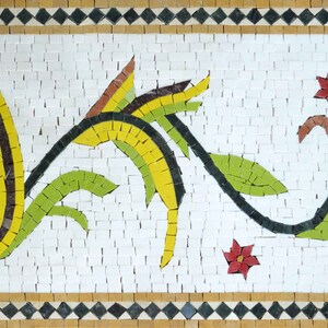 Könnte beinhalten: Mosaikkunstwerk mit floralem Design. Das Kunstwerk zeigt einen weißen Mittelgrund mit einer stilisierten Anordnung aus gelben, grünen und schwarzen Blättern und Stielen. Rote und blaue Blumen sind ebenfalls vorhanden. Der Rand ist eine Mischung aus braunen, schwarzen und weißen Fliesen.