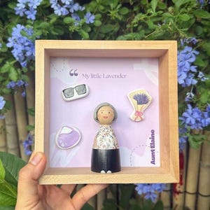 Può includere: Una cornice quadrata in legno contenente una bambolina con capelli grigi, un top a fiori e una gonna nera. Lo sfondo presenta il testo "My little Lavender" e "Aunt Elaine" con piccole icone di occhiali da sole, un bouquet di lavanda e una pochette viola.