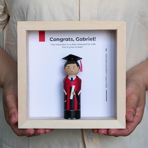 May include: A framed graduation gift for Gabriel, featuring a hand-painted wooden figure wearing a graduation cap and gown. The frame includes the text "Congrats, Gabriel!" and "Your education is a dress rehearsal for a life that is yours to lead." and "HARVARD UNIVERSITY Class of 2005".