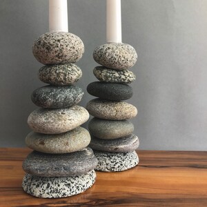 May include: Two candle holders made of stacked, smooth stones in shades of gray, tan, and black. Each holder has a white candle at the top. The stones vary in size and shape, creating a natural, organic look. The holders sit on a wooden surface.