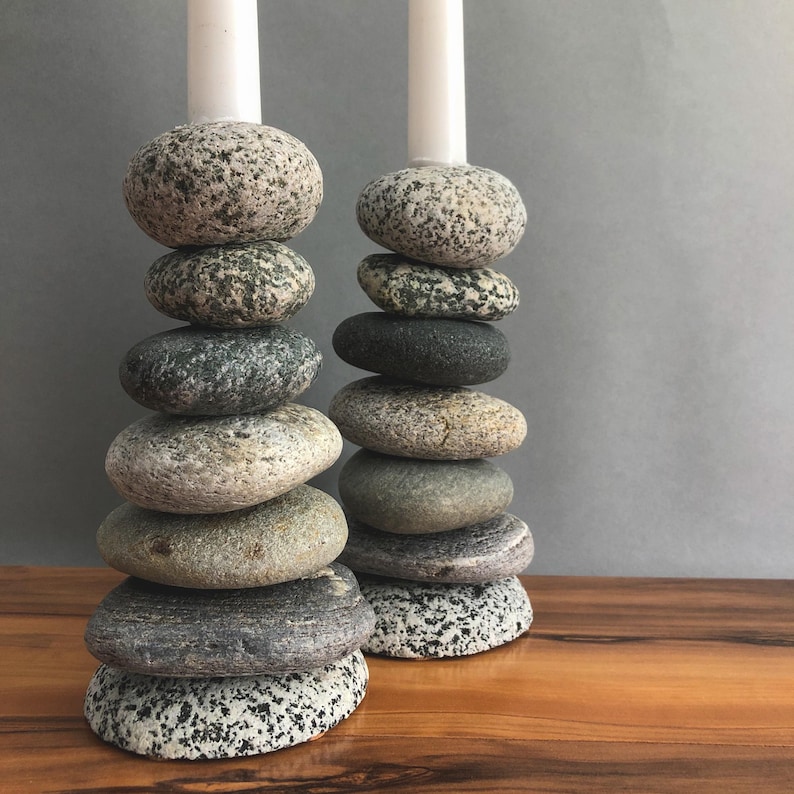 May include: Two candle holders made of stacked, smooth stones in shades of gray, tan, and black. Each holder has a white candle at the top. The stones vary in size and shape, creating a natural, organic look. The holders sit on a wooden surface.