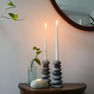 May include: Two lit white taper candles sit atop stacked stone candle holders. A small glass bottle with greenery and a lit stone candle are also on the wooden surface. A round mirror is in the background.