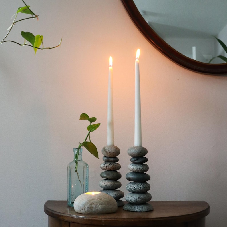 May include: Two lit white taper candles sit atop stacked stone candle holders. A small glass bottle with greenery and a lit stone candle are also on the wooden surface. A round mirror is in the background.