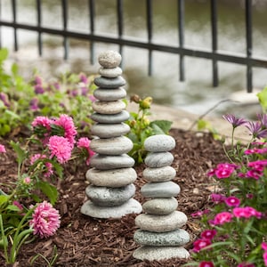 May include: Two stacks of balanced gray stones, resembling small towers, are set in a garden bed. The stones vary in size and shape. Pink flowers and green foliage surround the stone stacks, with a black metal fence in the background.