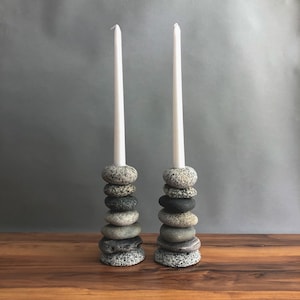 May include: Two candle holders made of stacked, smooth stones. Each holder supports a tall, white candle. The stones are various shades of gray and black, creating a natural, rustic aesthetic. The candles are white.