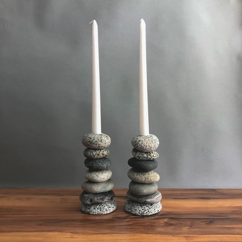 May include: Two candle holders made of stacked, smooth stones. Each holder supports a tall, white candle. The stones are various shades of gray and black, creating a natural, rustic aesthetic. The candles are white.