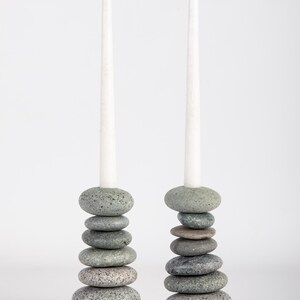 May include: Two stone candle holders, each composed of stacked, gray river stones. Each holder supports a tall, white taper candle. The stones vary in size and shade, creating a natural, textured appearance. The background is plain white.