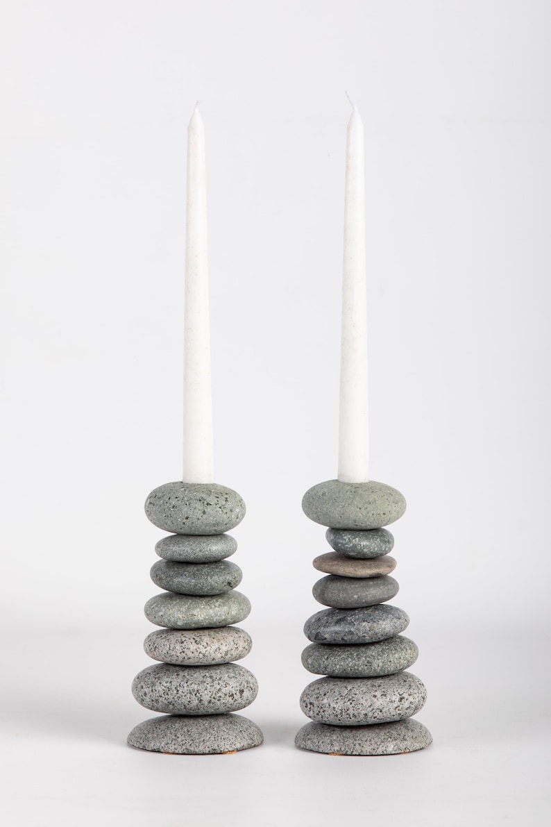 May include: Two stone candle holders, each composed of stacked, gray river stones. Each holder supports a tall, white taper candle. The stones vary in size and shade, creating a natural, textured appearance. The background is plain white.
